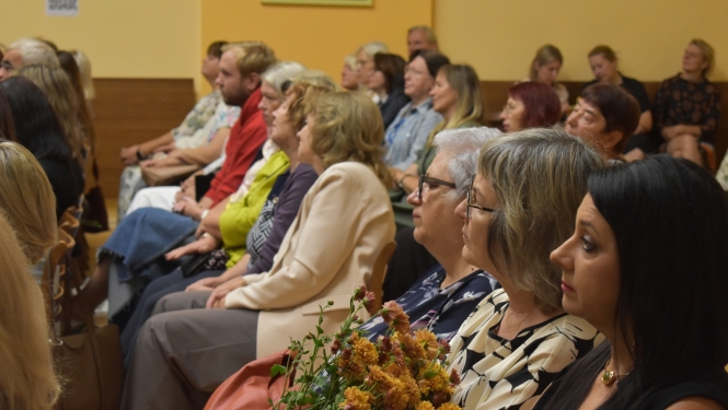 daudz cilvēku sēž zālē ar ziediem rokās