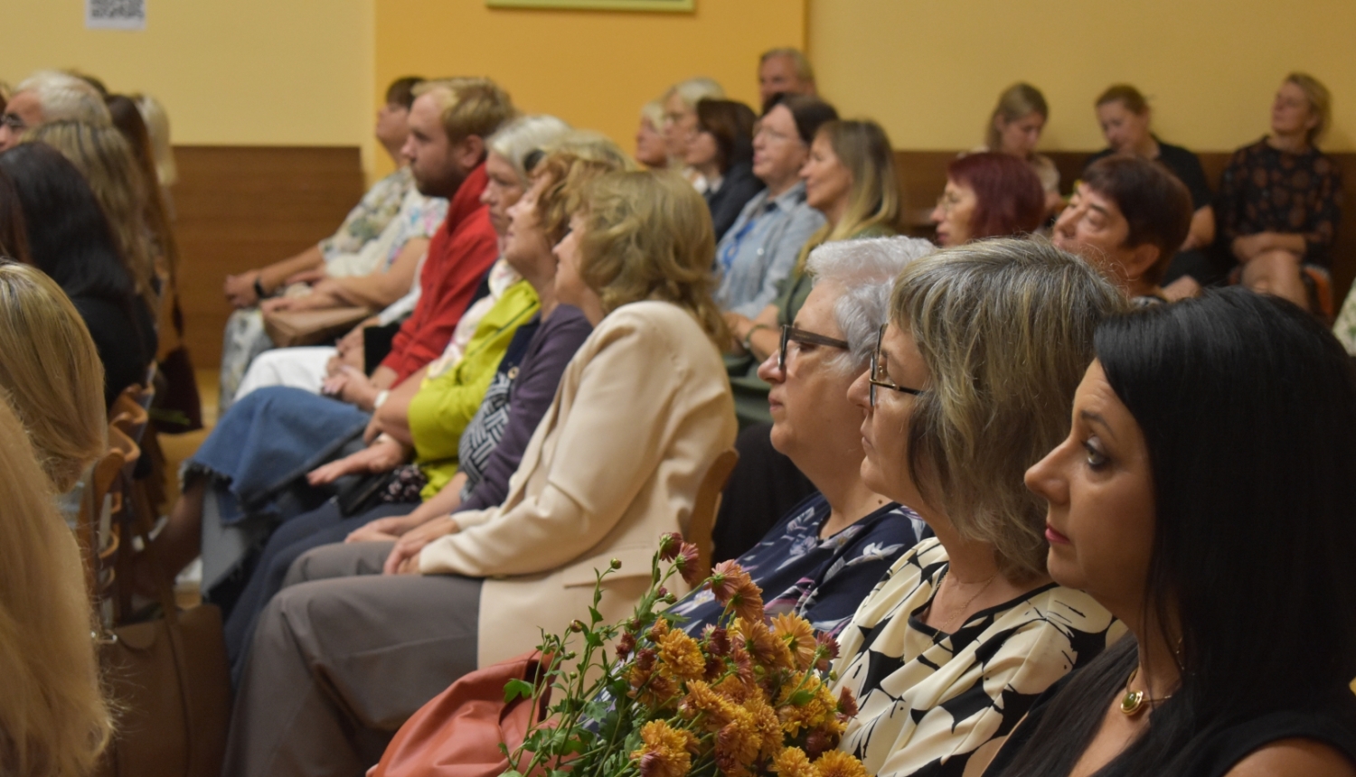 daudz cilvēku sēž zālē ar ziediem rokās