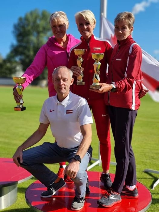 4 cilvēki (trīs sievietes sporta tērpos un vīrietis) uz goda pjedestālā, fonā trīs karogi