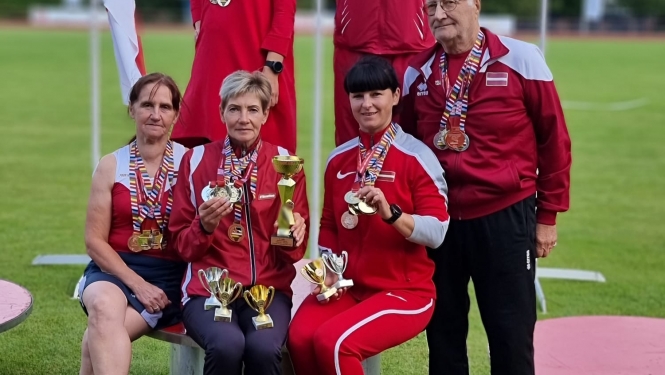 6 cilvēki sporta tērpos uz goda pjedestālā, fonā trīs karogi