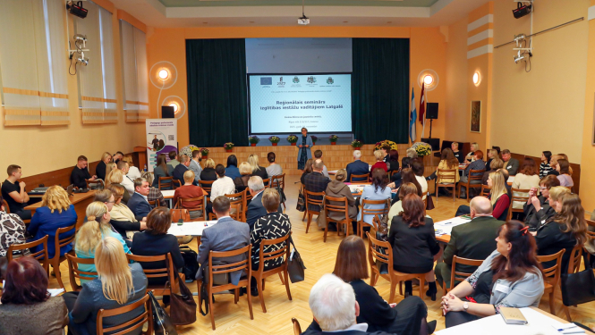 liela zāle pilna ar cilvēkiem, viens priekšā runā