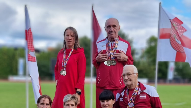 6 cilvēki sporta tērpos uz goda pjedestālā, fonā trīs karogi