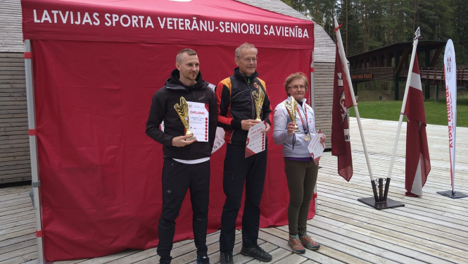 Divi vīrieši un sieviete stāv pie sarkanas telts ar diplomiem