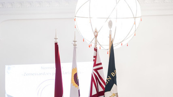 pie valsts karoga stāv sieviete un 3 vīrieši formas tērpos