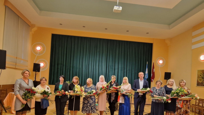 daudz cilvēku stāv rindā ar ziediem un diplomiem