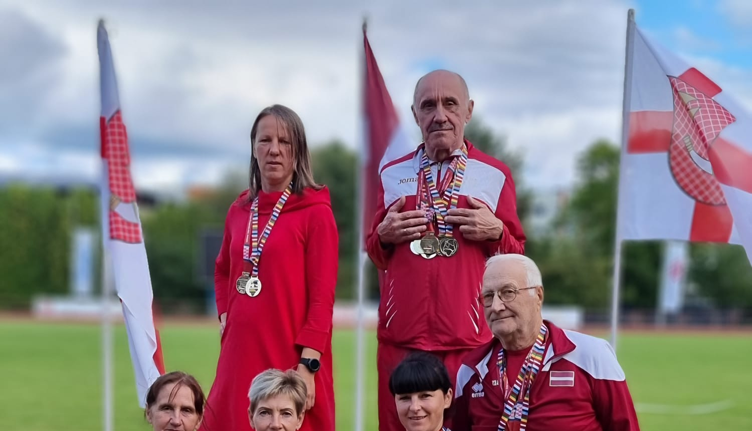 6 cilvēki sporta tērpos uz goda pjedestālā, fonā trīs karogi