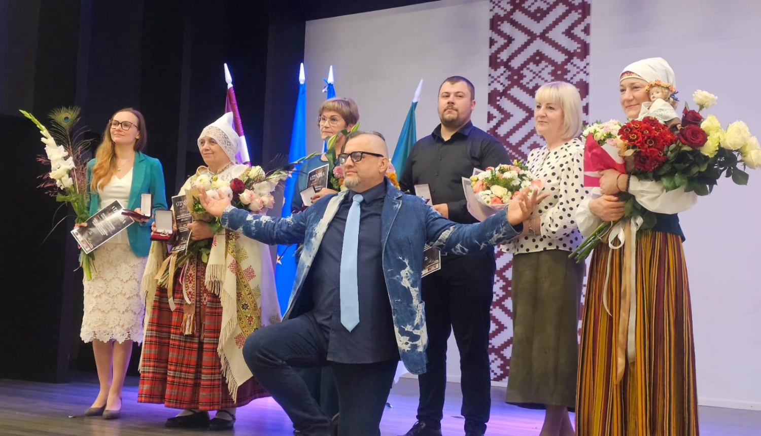 daudz cilvēki uz skatuves ar ziediem