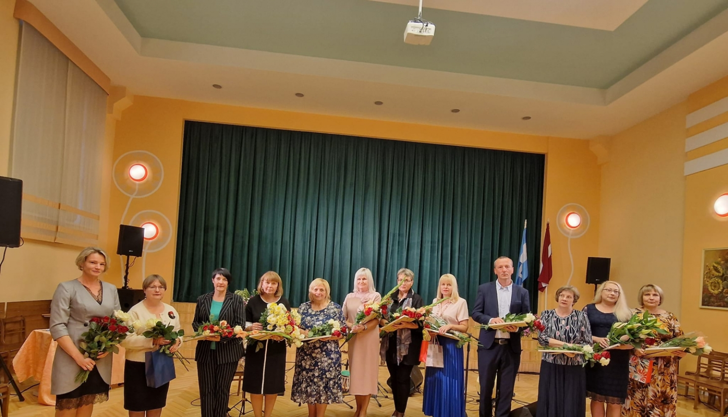 daudz cilvēku stāv rindā ar ziediem un diplomiem