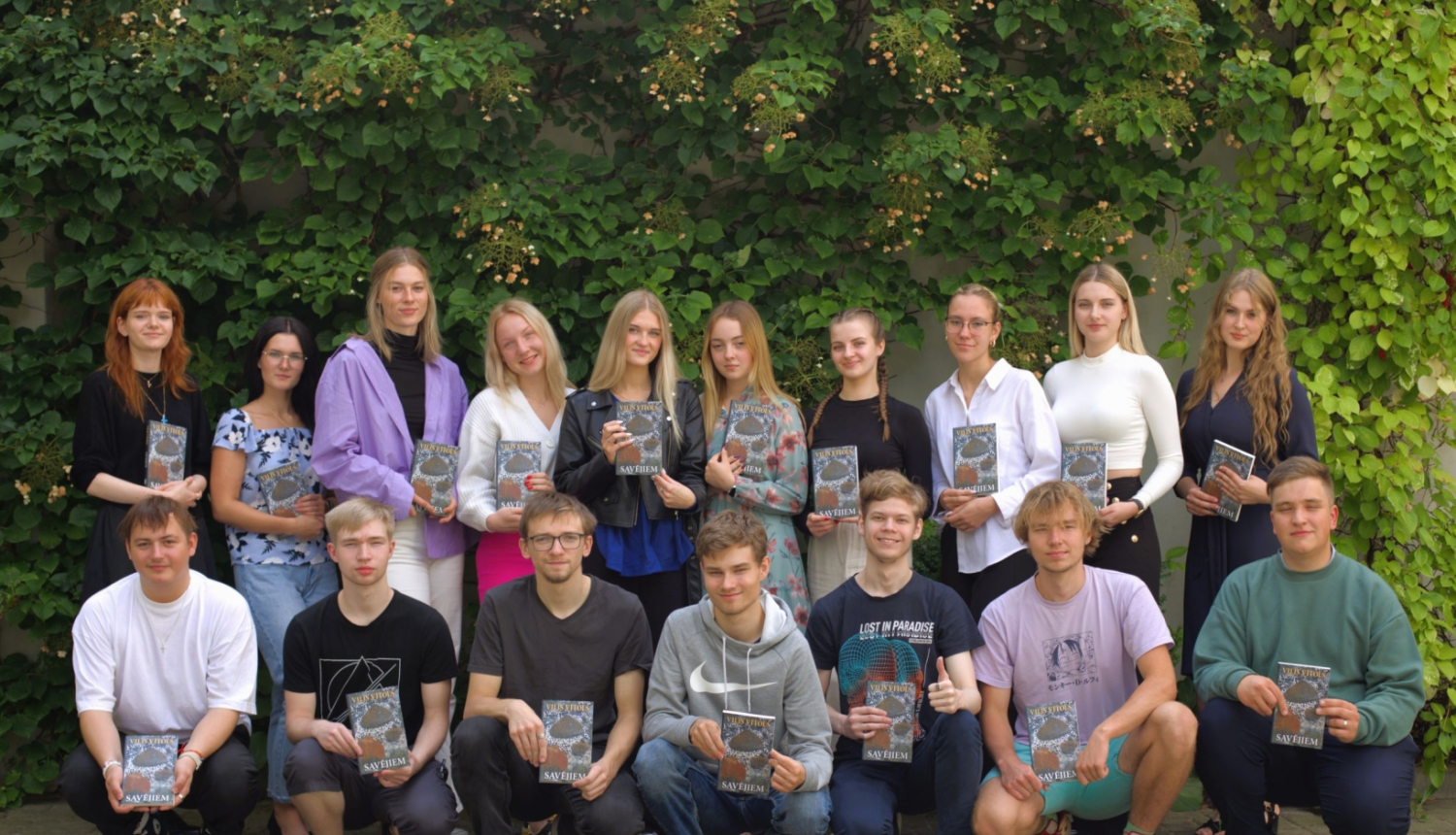 daudz jauniešu divās rindās pozē pie zaļas augu sienas