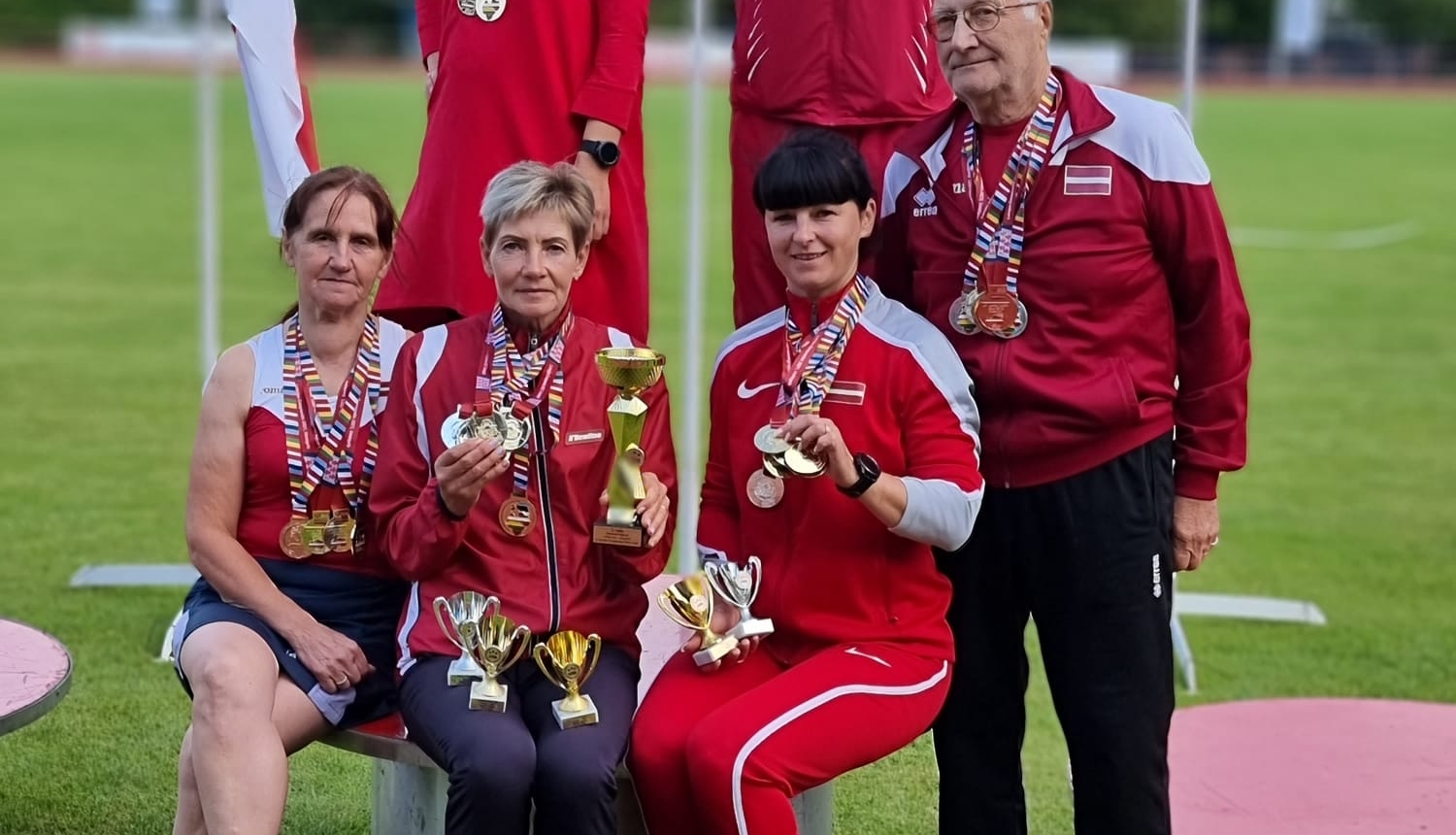 6 cilvēki sporta tērpos uz goda pjedestālā, fonā trīs karogi