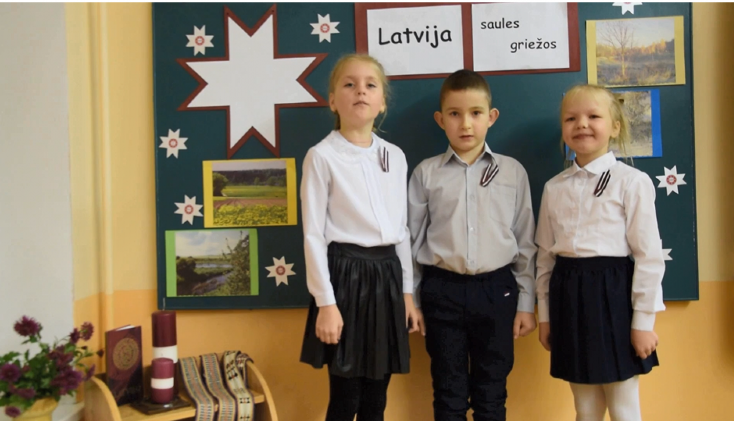 Trīs skolēni stāv klasē pie tāfeles