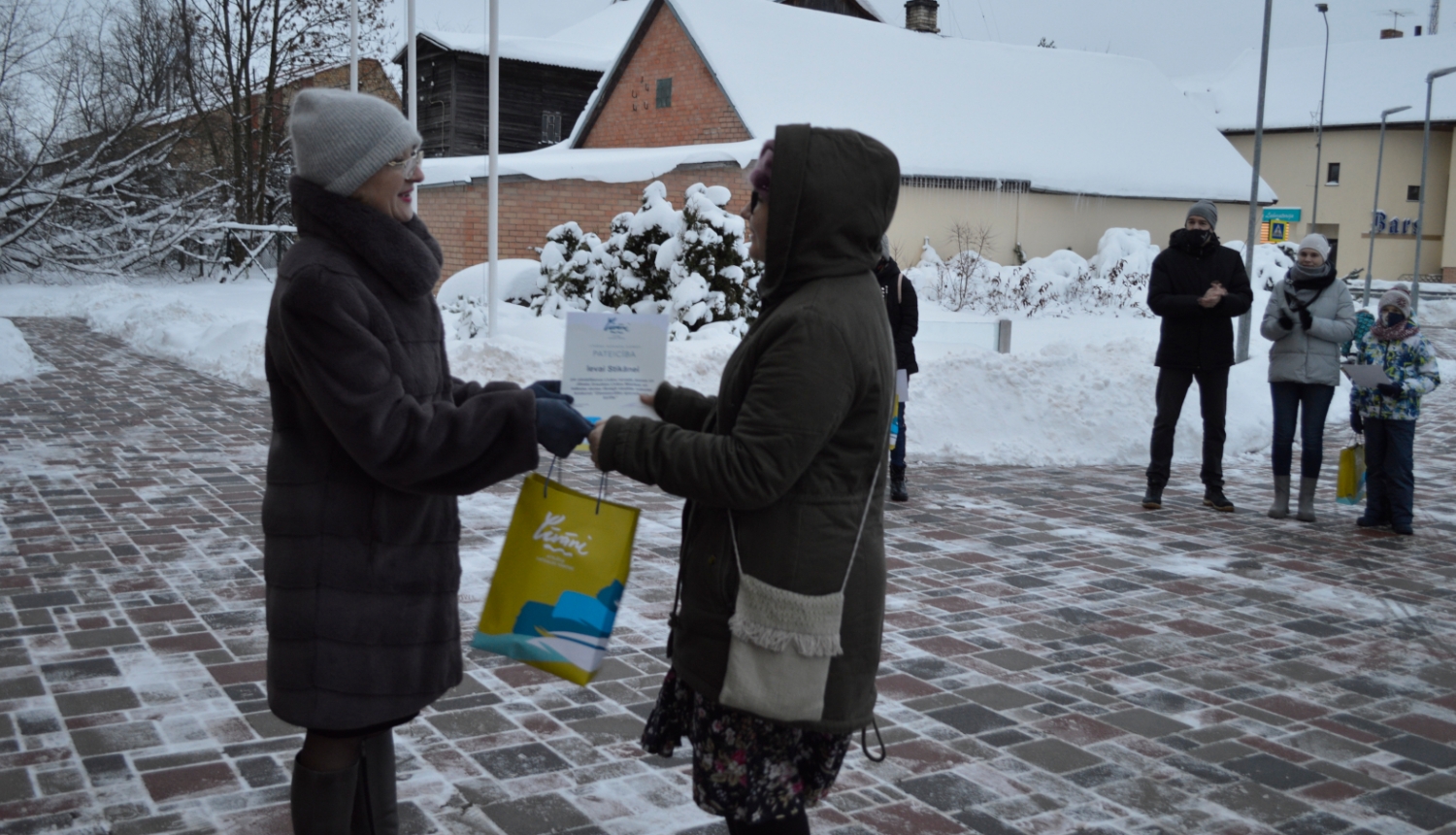 Sieviete meitenei pasniedz maisiņu un pateicības rakstu