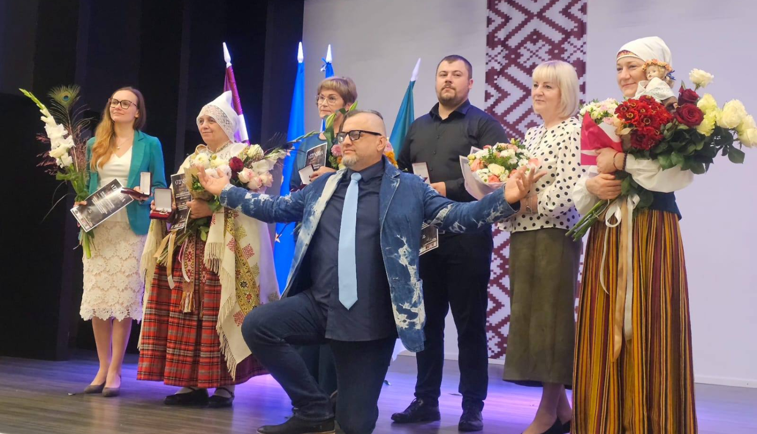 daudz cilvēki uz skatuves ar ziediem